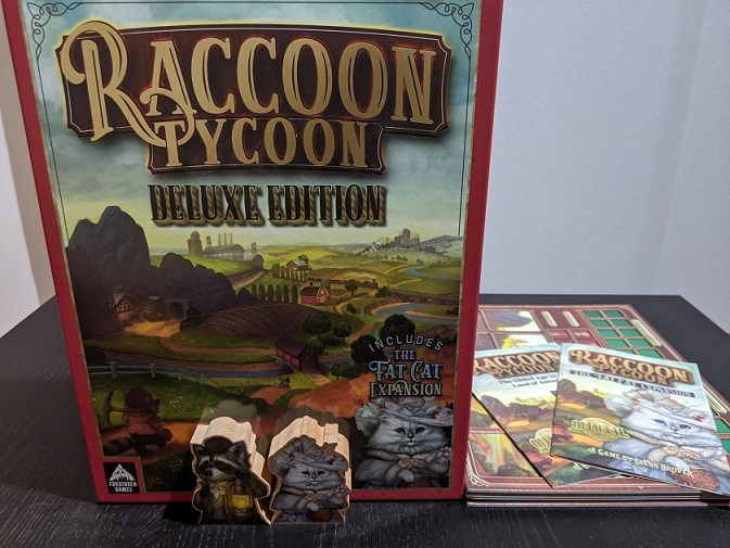 The Raccoon Tycoon and The Fat Cat Expansion first player tokens sit in front of the box. One is a Raccoon in a waistcoat with a lantern, the other is a fat fluffy white cat dressed in a Sunday dress clutching a purse.