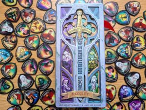 An ornate, silver, Excalibur-themed board game box is surrounded by dozens of colourful shield-shaped character tokens on a light wooden table. The box features an embossed central sword and six stained-glass windows depicting characters like Merlin and various knights.