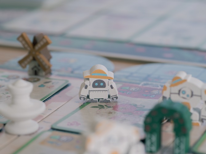 A macro shot highlighting custom screen-printed wooden game pieces. A white robot meeple wearing a straw hat takes center stage on a garden space, surrounded by a miniature brown windmill, a white fountain, and a green garden archway piece.