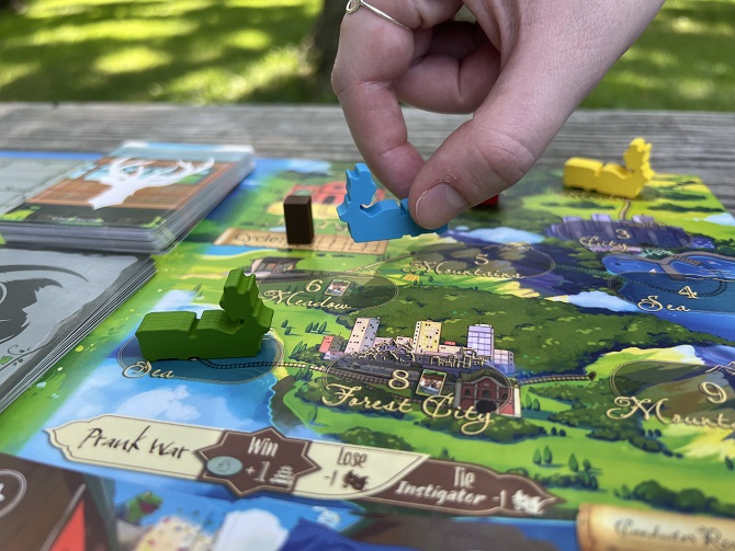 A close-up view of the Spirit Train game board during play, featuring illustrated region nodes such as Meadow, Forest City, Sea, and Mountains. A player's hand places a bright blue deer-shaped wooden marker onto the board near green and yellow player pieces and draw decks.
