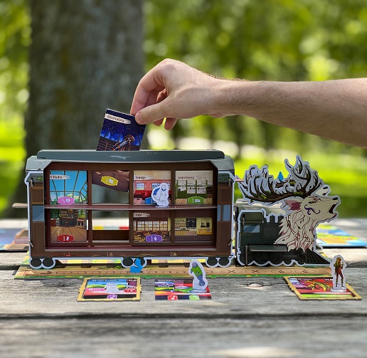 A close-up view of the 3D train carriage from Spirit Train resting on an outdoor wooden table. A hand inserts a square Observatory room card into the slotted roof of the two-story train model, which is fitted with a majestic stag-headed engine and surrounded by acrylic character standees and cards.