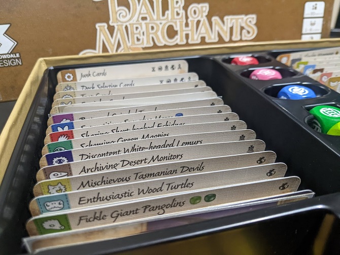 A close-up view inside the plastic insert of a Dale of Merchants box, featuring labelled card divider tabs for various animal suits like Fickle Giant Pangolins and Enthusiastic Wood Turtles next to custom dice.