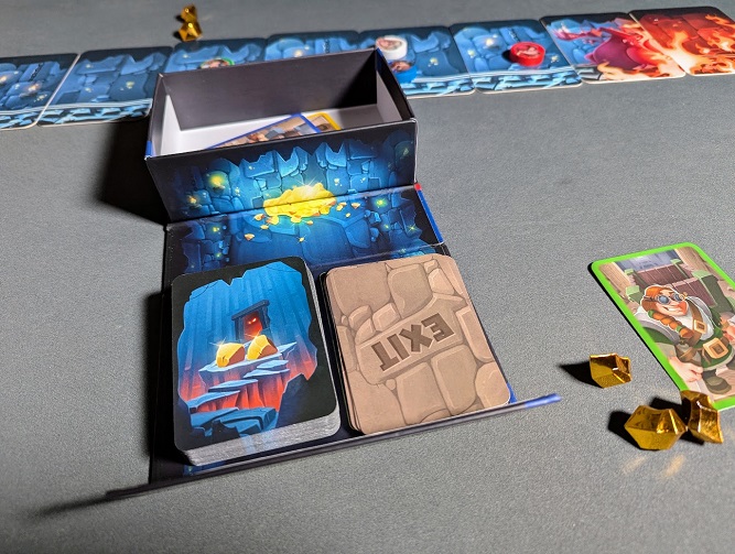 An open box for My Gold Mine unfolds to serve as a card holder for two draw piles: glowing gold nugget cards and brown Exit cards. Shiny gold plastic nuggets rest on the table beside a dwarf character card.