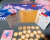 A trifold player aid card detailing player turns and scoring rules sits next to a pile of orange flower tiles. The background shows the game board with wooden tokens and a pink card featuring a hot air balloon.