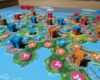 An angled shot of a Rebirth game in progress on a hex-grid map. Orange, blue, and pink plastic towers and castles are stacked and scattered across land tiles, surrounded by a sea of matching player tokens representing energy and food farms.